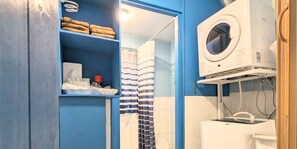 A bathroom with a blue wall, a white washing machine, a shelf with towels, and a striped shower curtain. - Avenue Koza Hotel (Okinawa)