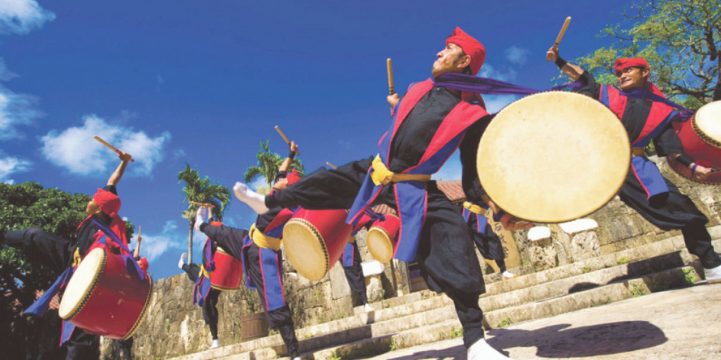 A group of drummers in traditional attire performing on stone steps with a clear blue sky in the background.