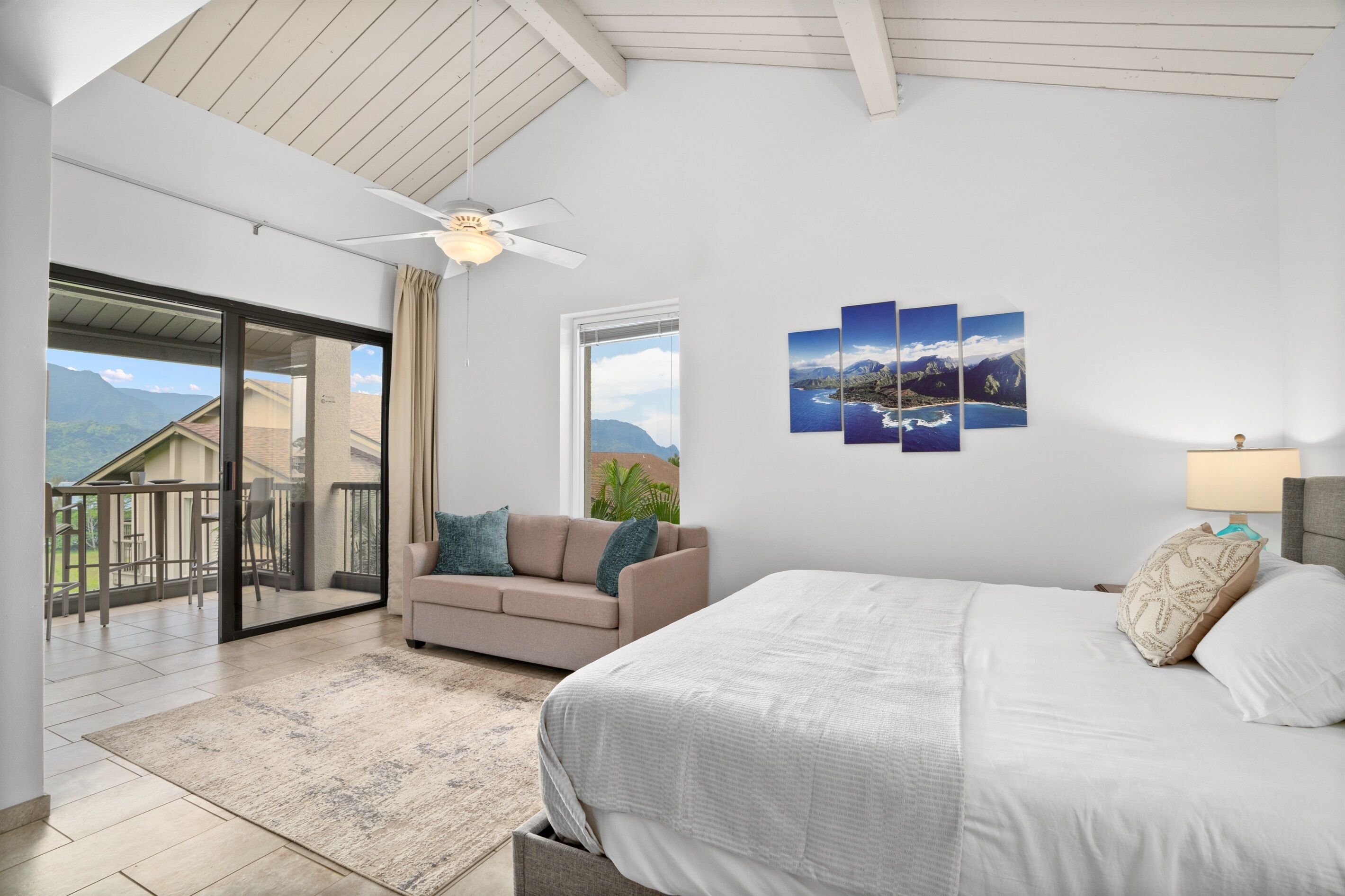 Un dormitorio con cama, sofá, ventilador de techo, ventana con vistas y puerta que da a un balcón.
