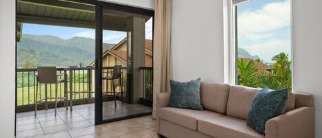 A living room with a beige sofa, two pillows, a large window, and a sliding glass door leading to a balcony with a view of mountains and a house.