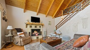 A cozy living room with a fireplace, a sofa, a coffee table, and a staircase. - Views! Sunsets! Amenities! A-Frame Cabin Sleeping 8 Guests. Come Relax & Unwind! (Lake Arrowhead)