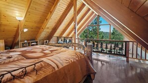 A bedroom with a sloped ceiling, a bed with a leaf-patterned bedspread, a nightstand, a lamp, and a window with a view of trees. - Views! Sunsets! Amenities! A-Frame Cabin Sleeping 8 Guests. Come Relax & Unwind! (Lake Arrowhead)
