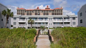 A multi-story hotel with a central courtyard, a wooden boardwalk leading to the entrance, and a red-roofed building in the background. - Oceanfront escape with pool & balcony views — peaceful 2BR retreat near Main Street & Cherry Grove (North Myrtle Beach)