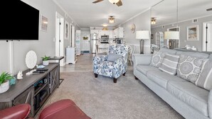 A spacious living room with a ceiling fan, a grey sofa, a patterned armchair, a red bench, and a kitchen in the background. - Oceanfront escape with pool & balcony views — peaceful 2BR retreat near Main Street & Cherry Grove (North Myrtle Beach)