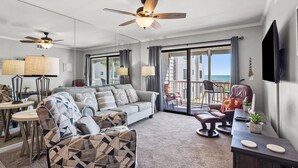 A spacious living room with a sofa, armchairs, a coffee table, and a TV. There are ceiling fans, a balcony with a view, and a dining area with chairs and a table. - Oceanfront escape with pool & balcony views — peaceful 2BR retreat near Main Street & Cherry Grove (North Myrtle Beach)