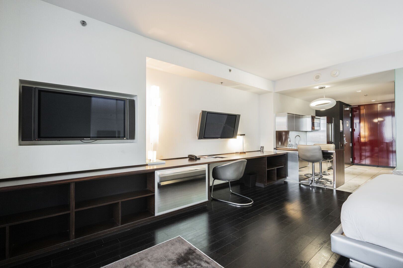 A modern hotel room with a large bed, a desk with a chair, a TV mounted on the wall, and a separate dining area.