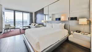 Moderni hotellihuone, jossa on suuri sänky, oleskelualue sohvineen ja nojatuoleineen, työpöytä sekä suuri ikkuna, josta avautuu näkymä kaupunkiin.