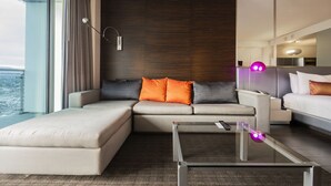 Moderni hotellihuone, jossa on suuri ikkuna, oransseilla ja harmailla koristetyynyillä varustettu sohva, lasinen sohvapöytä ja valkoisilla lakanoiden kanssa varustettu sänky.
