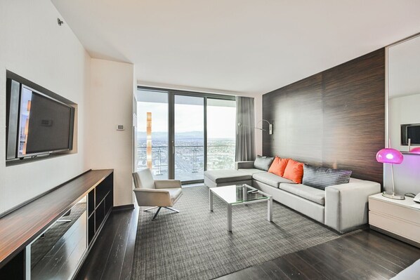 A modern hotel room with a large window offering a city view, a flat-screen TV mounted on the wall, a sofa with orange cushions, and a small table with a lamp. - Studio • Balcony • No Resort Fee • Free Parking • WO51 (Las Vegas)