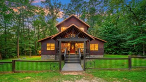 A log cabin with a porch and steps leading to the entrance, surrounded by a fenced yard and dense forest. - Rustic Family Getaway 2BR - Hot Tub, Fire Pit, Pool Table, Pets OK (Broken Bow)