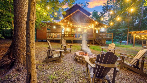A wooden cabin with a deck, surrounded by Adirondack chairs and a fire pit, set in a wooded area. - Rustic Family Getaway 2BR - Hot Tub, Fire Pit, Pool Table, Pets OK (Broken Bow)