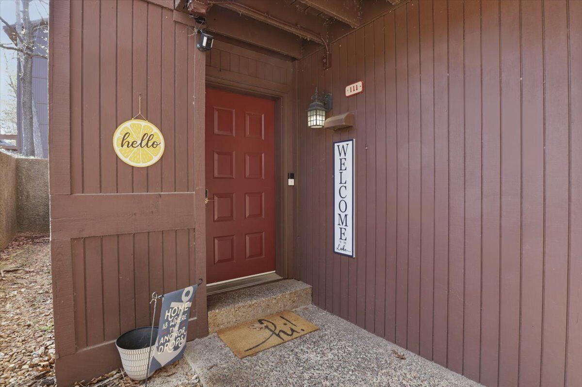Un bâtiment en bois avec une porte rouge, une pancarte indiquant « bonjour », une autre indiquant « bienvenue » et un paillasson orné d’une empreinte de main.