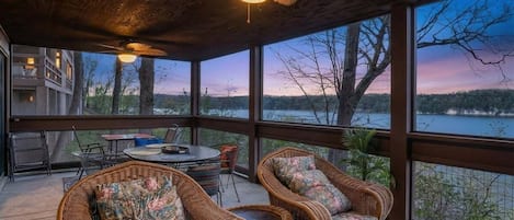 A covered outdoor seating area with wicker furniture and a ceiling fan, overlooking a body of water and trees.