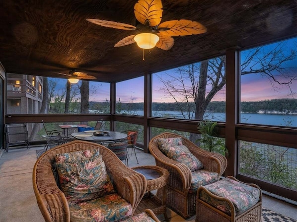 A covered outdoor seating area with wicker furniture and a ceiling fan, overlooking a body of water and trees.