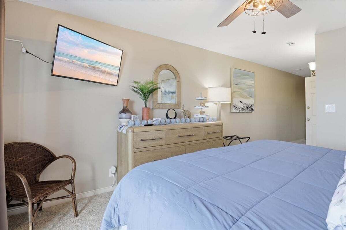 Um quarto com cama, cômoda, cadeira, televisão fixada na parede e ventilador de teto.