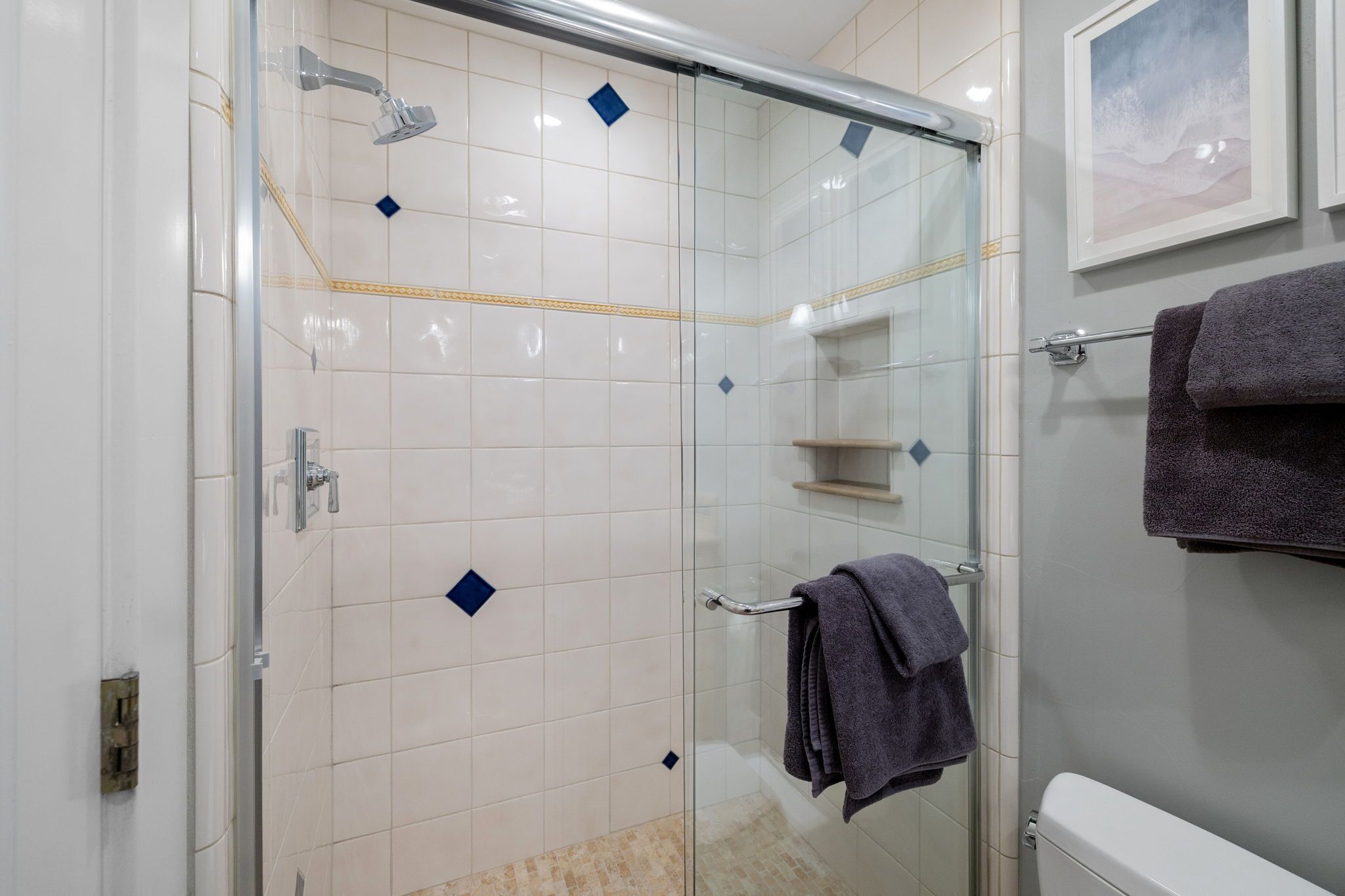 Une salle de bain moderne avec une douche à parois de verre, des carreaux blancs rehaussés d’accents bleus, des toilettes et un porte-serviettes avec des serviettes.