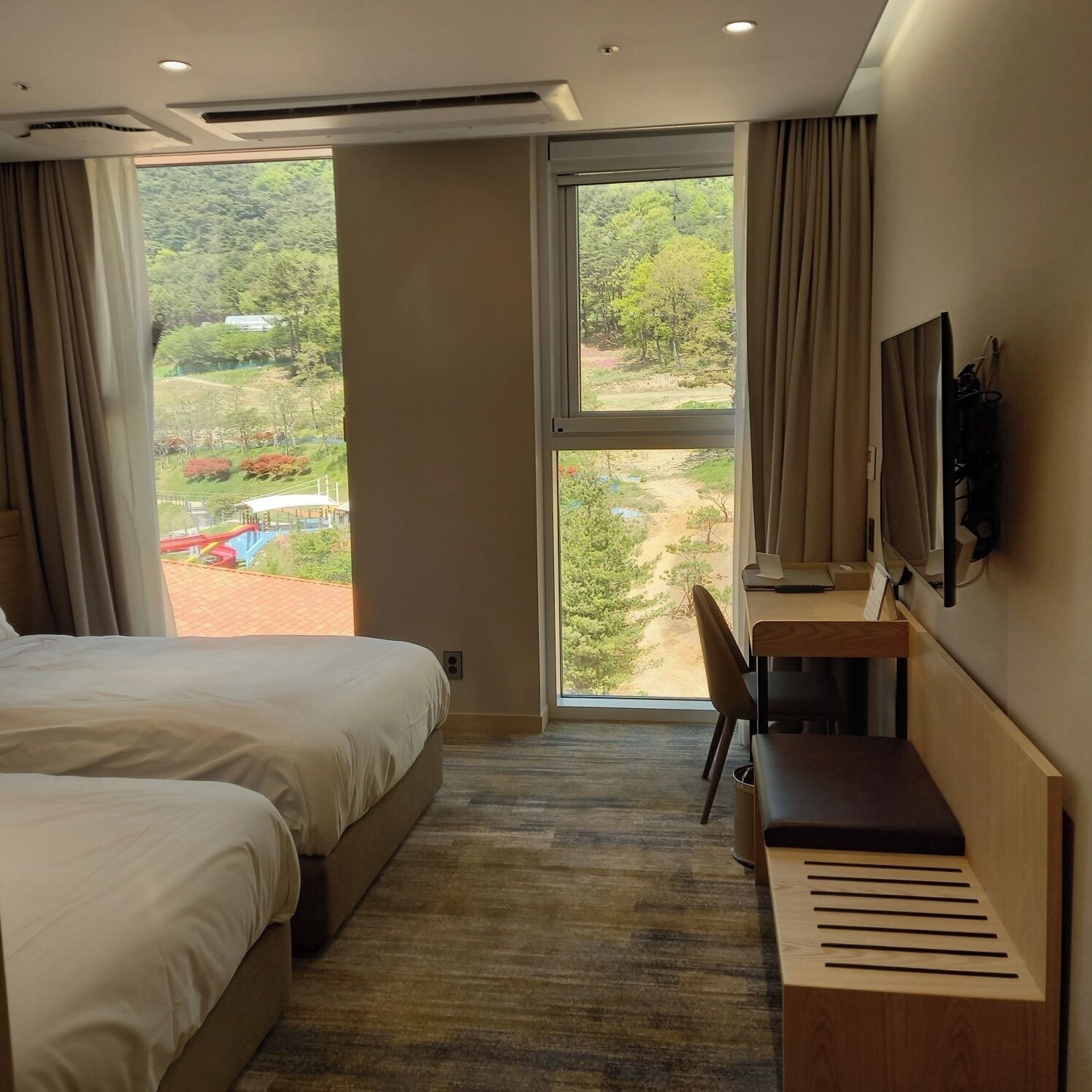 A hotel room with two beds, a desk, a chair, a TV mounted on the wall, and a large window offering a view of a landscaped area.