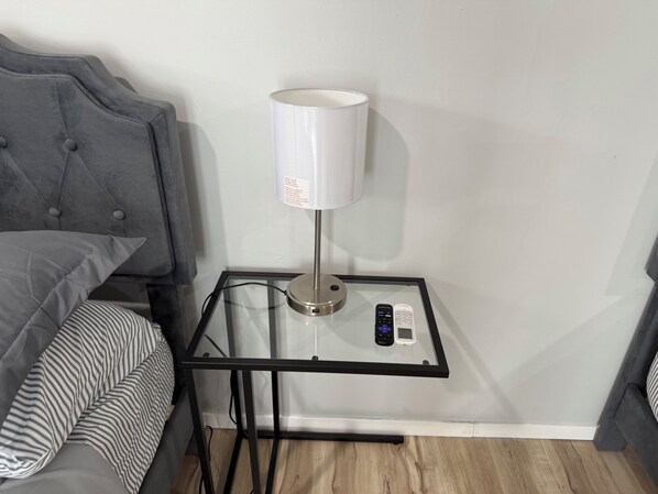 A modern hotel room with a grey tufted headboard, a glass side table with a white lamp, a remote control, and a smartphone.