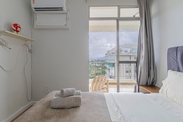 A bedroom with a bed, a window with a view of buildings, a ceiling fan, and a wall-mounted air conditioner.