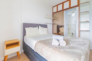 A neatly made bed with a grey headboard, a bedside table, and a mirror with shelves above it.