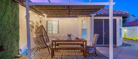 An outdoor patio with a wooden table, chairs, and a pergola with string lights.