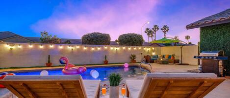 Un área de piscina con sillones de descanso, una parrilla y luces colgantes.