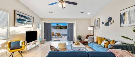 A modern living room with a blue sofa, a TV, and a ceiling fan. There is a door leading to an outdoor area with a view of the sky.