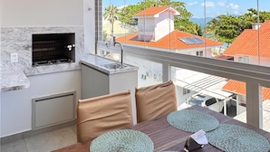 A modern kitchen with a built-in grill, a dining area with a wooden table and green placemats, and a large window offering a view of red-roofed houses and greenery. - Cachoeira do bom jesus  (Florianopolis)