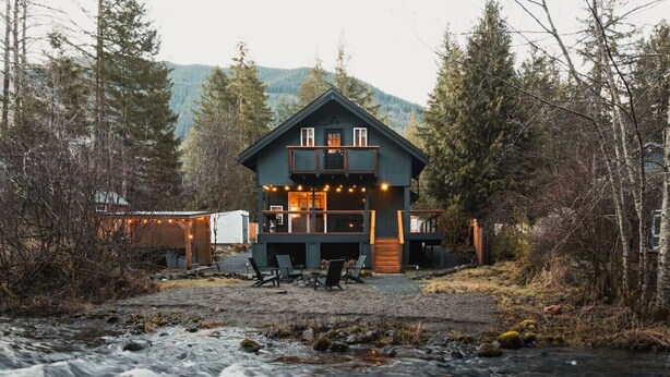 Une cabane moderne à deux étages, dotée d’un balcon, située au bord d’une rivière et entourée d’une forêt dense et de montagnes.