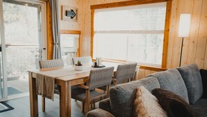 Une pièce chaleureuse avec une table à manger en bois, des chaises en osier, un canapé gris et de grandes fenêtres.