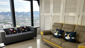 A modern living room with a grey sofa and a beige sofa with yellow trim, both with decorative pillows. A large window offers a city view. - Food Paradise Georgetown 218 Macalister 2BR Family Suite By Smart Space Home (George Town)