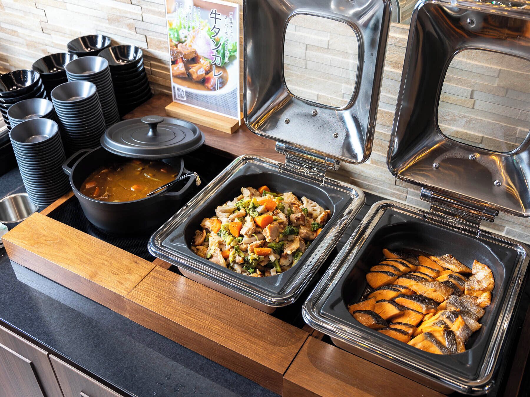 Un buffet proposant divers plats, notamment un pot de soupe, un plat de poulet et de légumes, et un autre de poisson grillé.