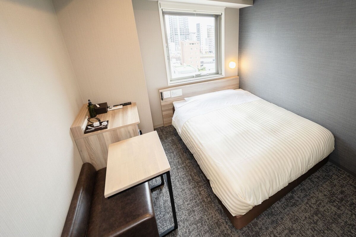 Une petite chambre d’hôtel comprenant un lit, un bureau, une chaise, une fenêtre donnant sur la ville et une lampe fixée au mur.