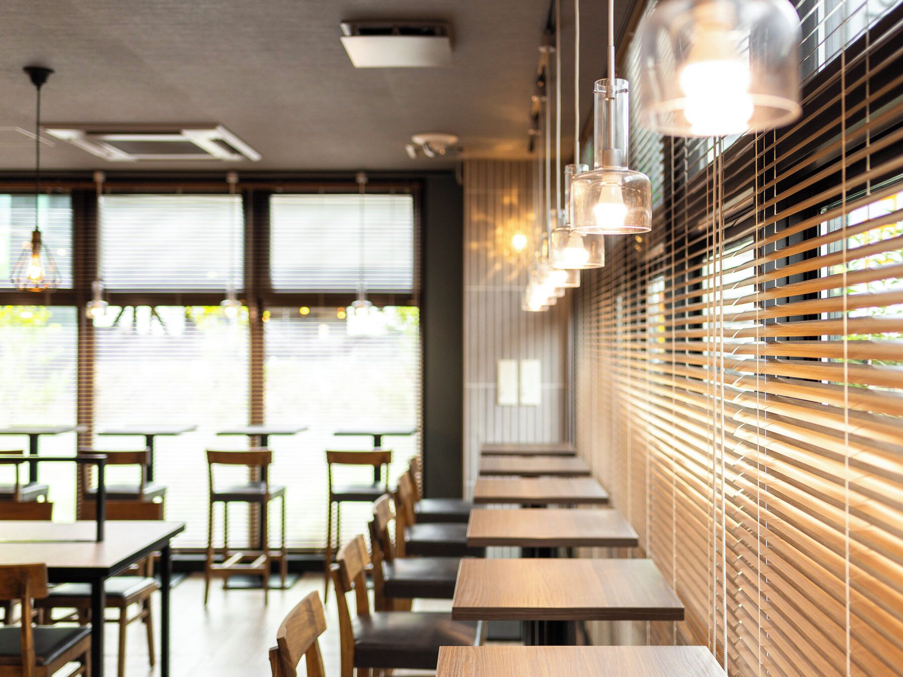 L’intérieur d’un restaurant avec des tables et des chaises en bois, de grandes fenêtres équipées de stores et des lampes suspendues.