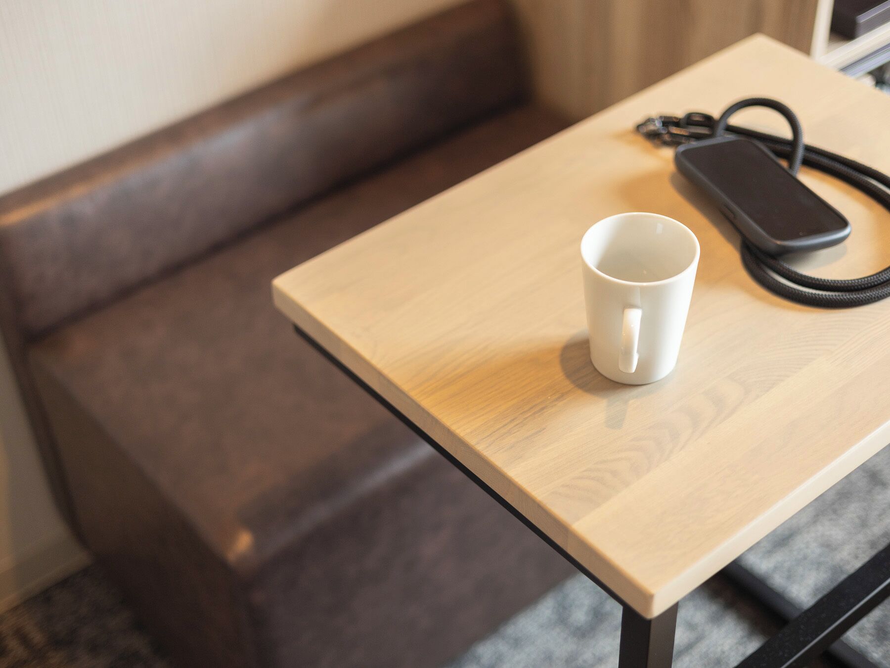 A wooden table with a white mug and a black phone charger.