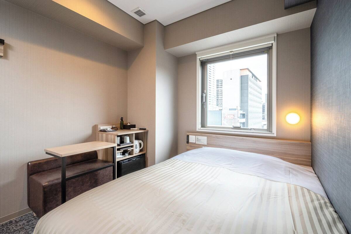 Une chambre d’hôtel comprenant un lit, un bureau avec une bouilloire, un petit coin salon et une fenêtre donnant sur la ville.