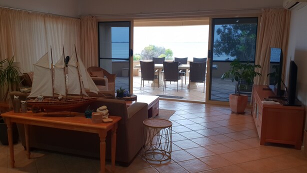 Un salón con una mesa de madera, un sofá, un taburete pequeño y redondo, y un modelo de barco. Hay una puerta corrediza de vidrio que da a un patio exterior con una mesa y sillas.