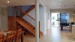 Un comedor amplio con una escalera de madera que lleva a un nivel superior, una mesa de billar en la habitación contigua y vistas al exterior a través de amplios ventanales.
