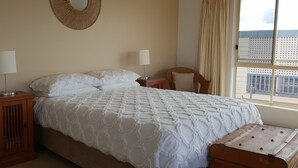 Una cama bien hecha con colcha blanca, dos lámparas de noche, una silla de mimbre y vistas de un edificio a través de la ventana.
