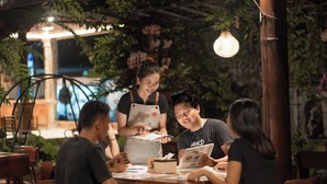 A group of people seated at a wooden table in an outdoor setting with greenery and hanging lights. - The central lovina hotel & restaurant (Buleleng)