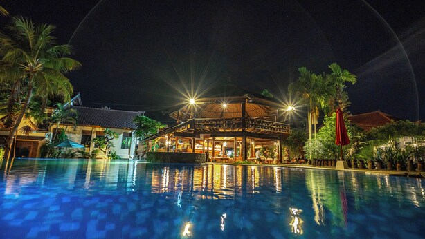A nighttime view of a hotel with a swimming pool, illuminated by street lamps, surrounded by palm trees and traditional architecture. - The central lovina hotel & restaurant (Buleleng)
