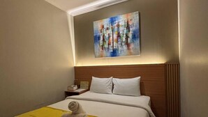 A hotel room with a neatly made bed, a headboard, a bedside table with a lamp, and a painting above the bed. - The central lovina hotel & restaurant (Buleleng)