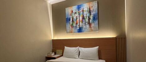 Una habitación de hotel con una cama bien hecha, cabecera, mesita de noche con lámpara y un cuadro sobre la cama.