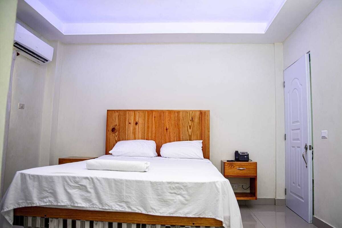 Une chambre à coucher avec une tête de lit en bois, un lit recouvert de draps blancs, une table de chevet avec un téléphone et une porte.