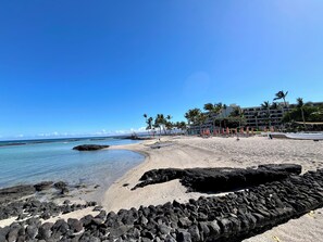 A beach with a rocky foreground, a clear blue sky, and a hotel with multiple floors and palm trees in the background.