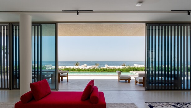 Una sala de estar moderna con un sofá rojo, puertas de cristal grandes que dan a una zona junto a la piscina con mobiliario exterior.