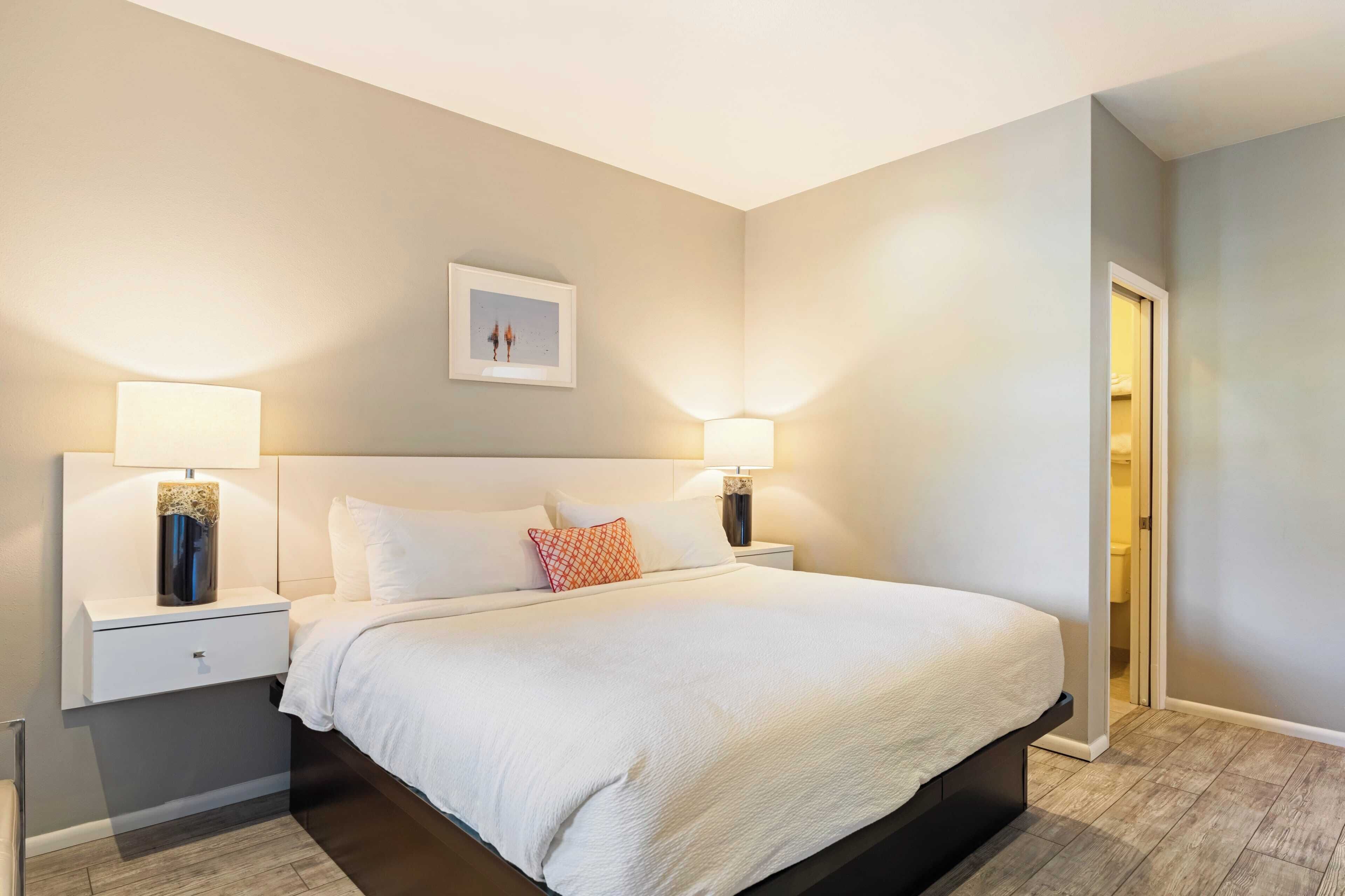 A neatly arranged bedroom with a large bed, two bedside tables with lamps, a small artwork on the wall, and a door leading to a bathroom.