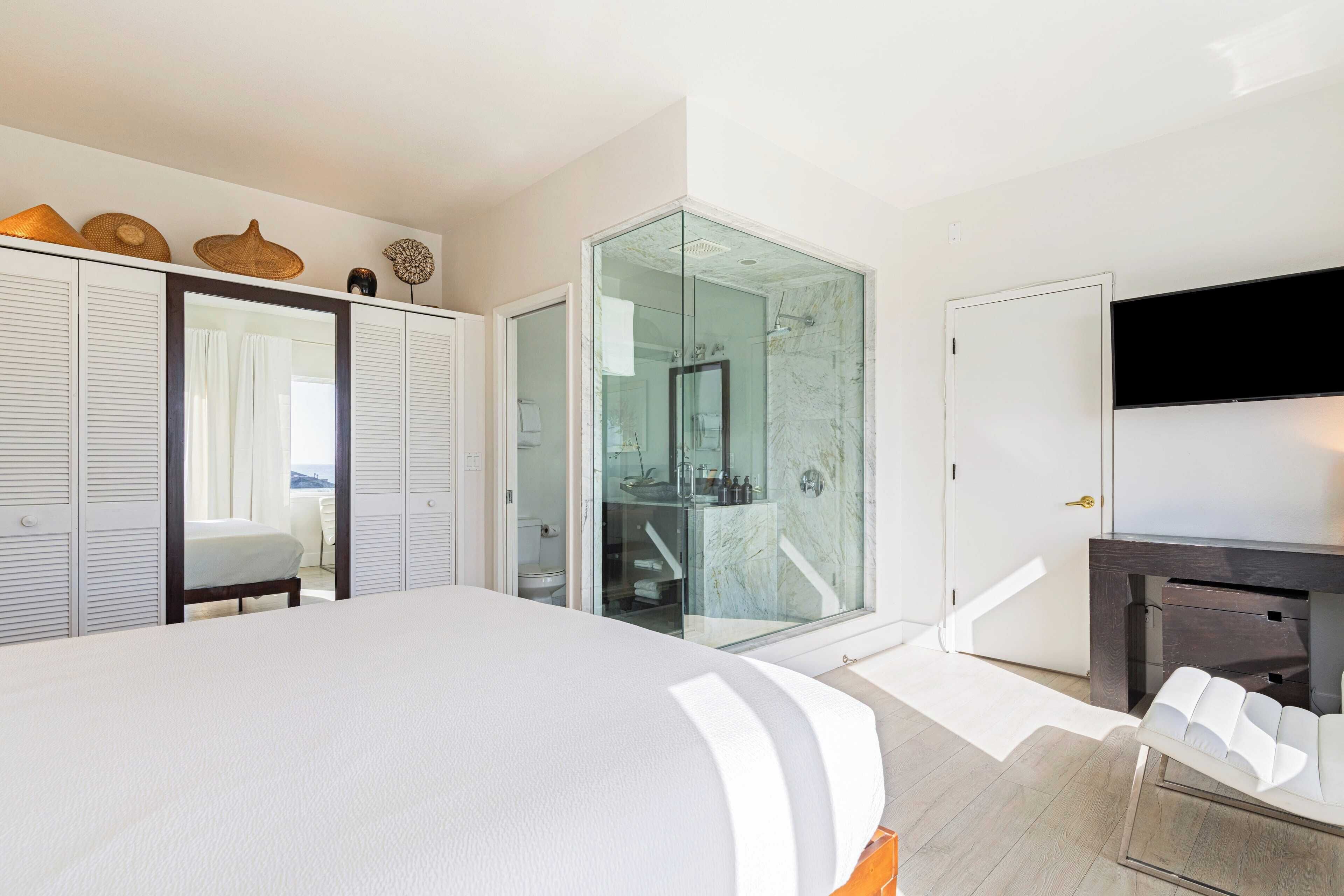 Ein modernes Hotelzimmer mit einem großen Bett, einer gläsernen Dusche, einem an der Wand montierten Fernseher und einem Schrank mit Schiebetüren.