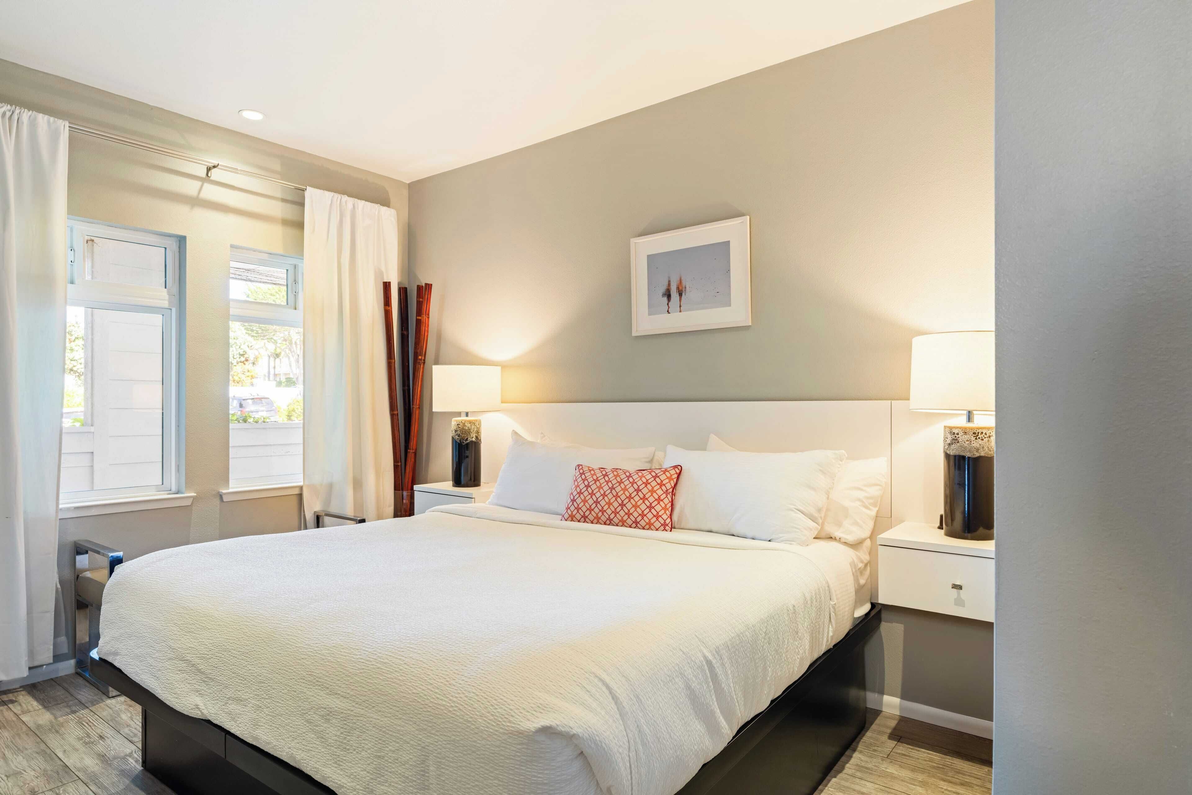 A neatly arranged bedroom with a large bed, two bedside tables with lamps, a framed picture on the wall, and a window with curtains.