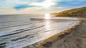 A beach with a wooden pier extending into the water, a sandy shore, and a hilly landscape in the background.